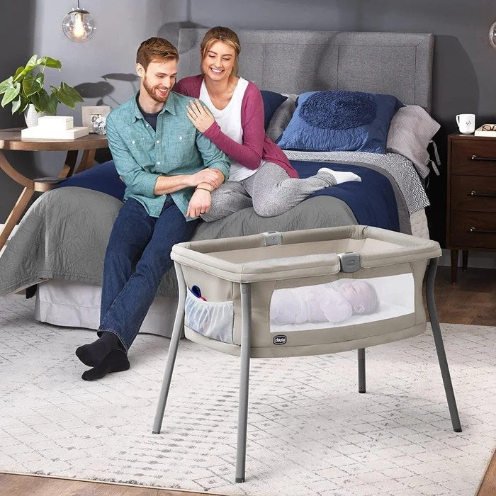 Parents smiling at their baby asleep in a Chicco LullaGo Anywhere bassinet