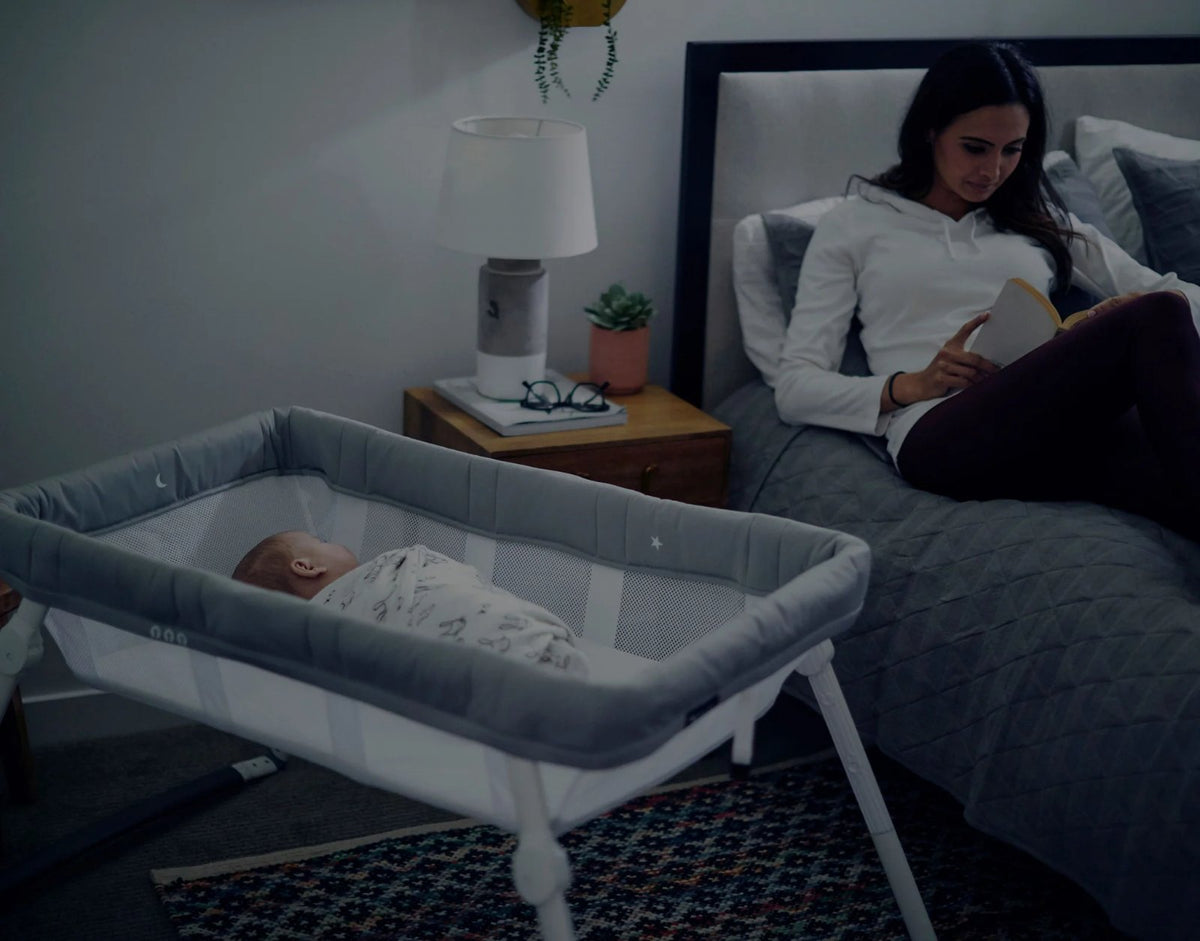 Mother reading in bed beside her baby asleep in a Guava Lotus Travel Bassinet