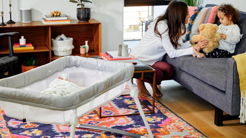 Baby asleep in a Guava Lotus Travel Bassinet while mum plays with her toddler