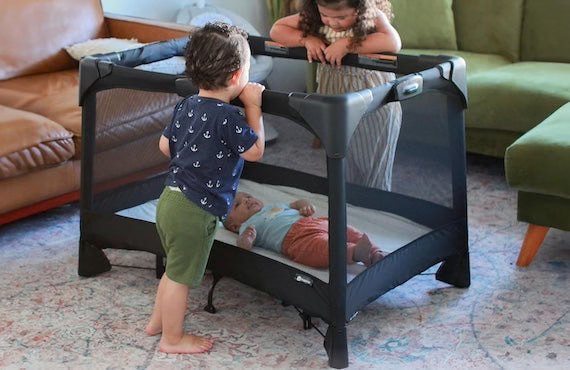Two toddlers looking at a baby in a 4moms Breeze playard