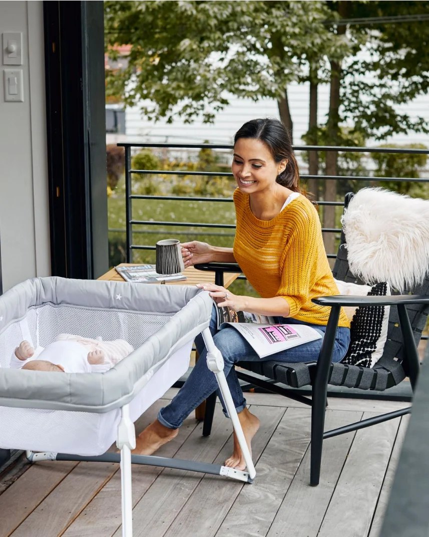 Mother on a deck smiling at her baby in a Guava Lotus Travel Bassinet
